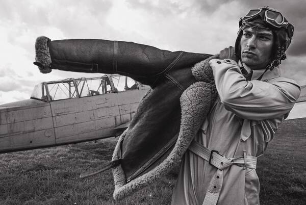 Ein junger Mann in historischer Fliegermontur zieht sich eine Lammfelljacke an. Im Hintergrund steht ein historisches Flugzeug. Schwarz-Weiß.