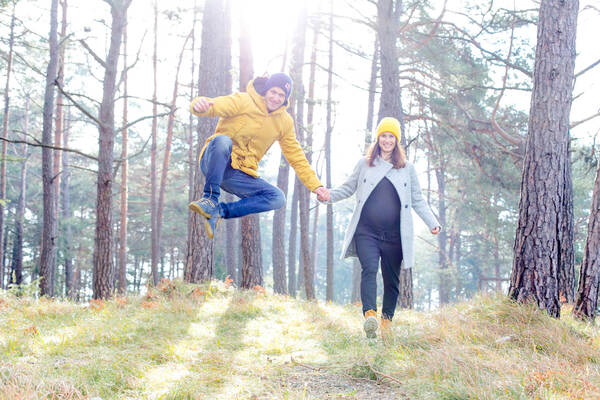 Ein Paar in blau und gelb kekleidet in einem Wald. Die Frau ist schwanger, der Mann macht einen Luftsprung.
