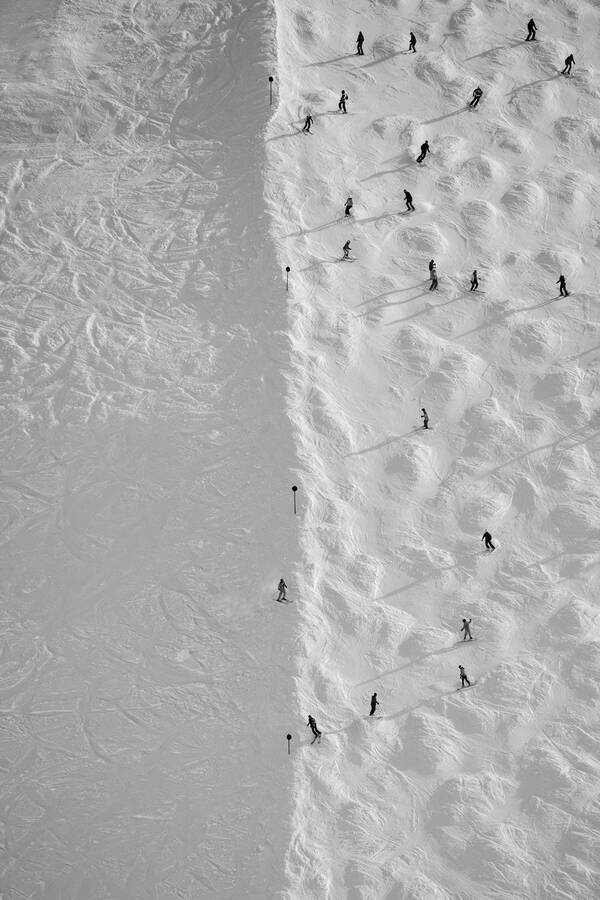 Blick von oben auf einen verschneiten Berghang. durch die Mitte des Bildes verläuft von oben nach unten die Begrenzung der Schipiste. rechts davon sind viele Schifahrer, links davon keine. Schwarz-Weiß.