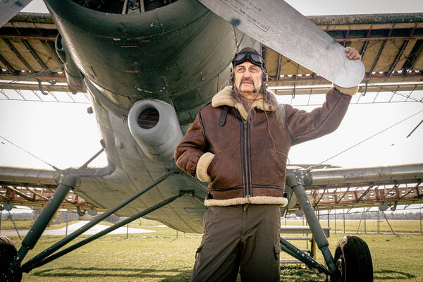 Ein Mann mit ausdrucksstarkem Schnauzbart in Fliegerjacke und historischem, ledernem Fliegerhelm mit Brille steht vor einem historischen Flugzeug, eine Hand am Propeller, die andere in die Lederjacke eingesteckt.