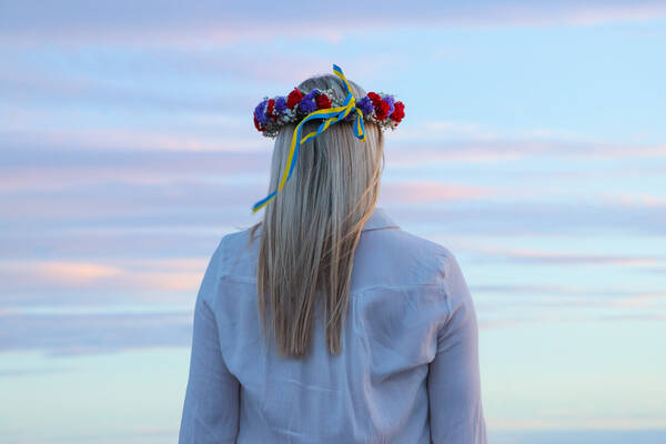 Eine junge, blonde Frau mit einem Blumenkranz am Kopf von hinten betrachtet vor einem pastellfarbigen Abendhimmel.