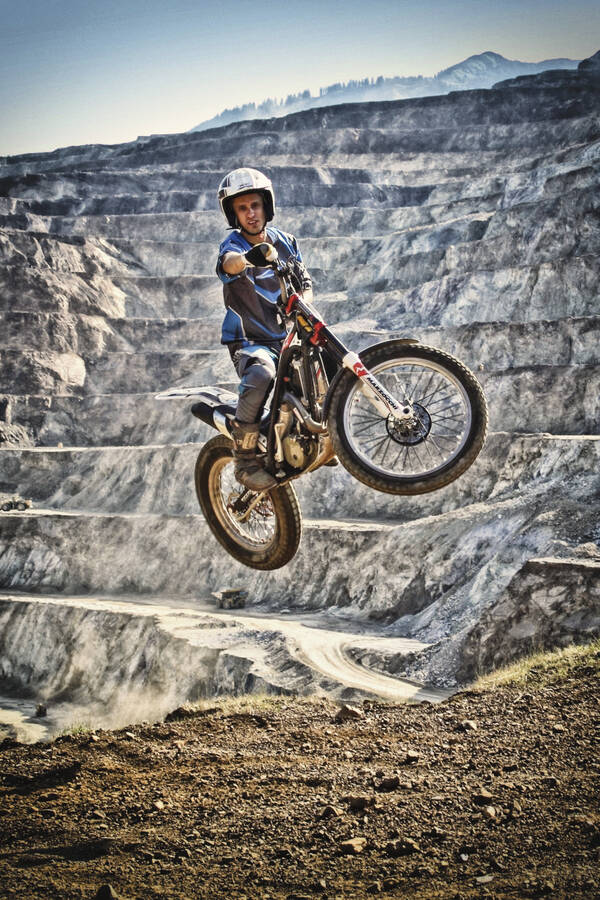 Bild eines Motocross-Fahrers der mit seiner Maschine gerade im Sprung ist. Im Hintergrund die vom Bergbau geschaffenen Hangstufen des steirischen Erzberges.