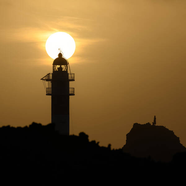 Abendstimmung. Ein Leuchtturm berührt die Untergehende Sonne. Auf einem Felsen neben dem Leuchtturm ist die Silhouette eines Menschen zu sehen.