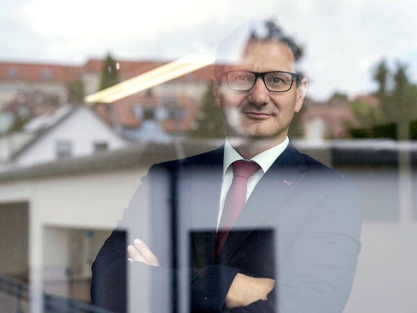 Portrait eines Mannes mit Brille in dunklem Anzug mit dunkelroter Krawatte und verschränkten Armen durch eine Fensterscheibe, in der sich die umgebenen Häuser spiegeln.