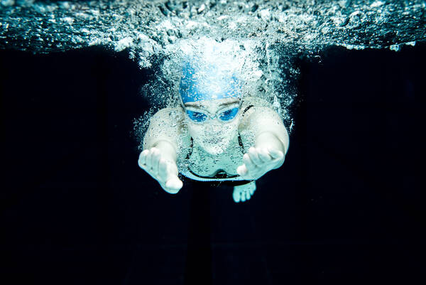 Eine Schwimmerin mit Schwimmbrille und Badehaube unter Wasser von vorne fotografiert.