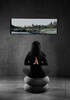 Eine Frau sitzt im Yogasitz mit hinter dem Rücken gefalteten Händen auf zwei übereinandergestapelten grauen Sitzkissen in einem betongrauen Raum und schaut auf ein Panoramabild einer unberührten Uferlandschaft.