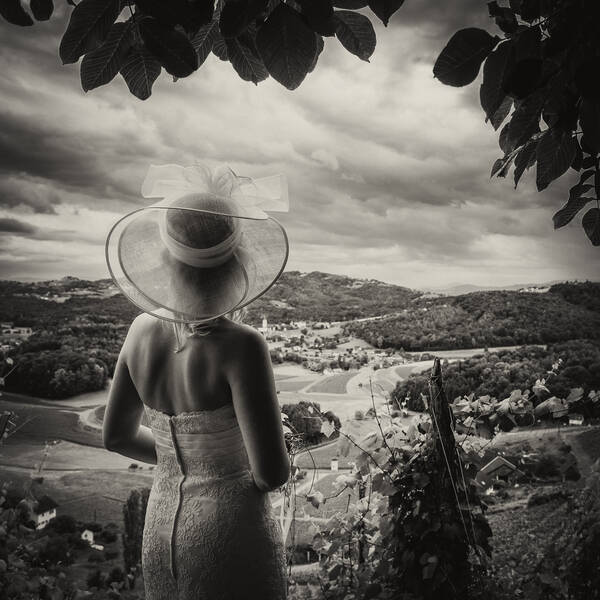 Portrait einer Braut mit schulterfreiem Kleid und transparentem Strohhut, die vom Weingut Harkamp in die Landschaft schaut. Duoton. Schwarz-Weiß.