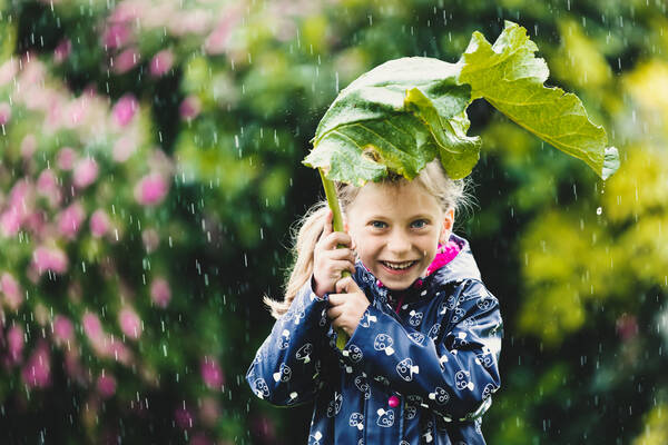 Ein Mädchen hält sich im Regen ein großes Blatt wie einen Schirm über den Kopf.