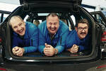 Drei Männer mit Schnauzbart in blauen Arbeitsanzügen liegen eng zusammengedrückt im Kofferraum eines Autos und lachen in die Kamera.
