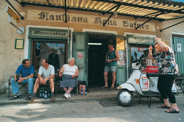 Ladenfront eines alten Kaufladens. Über der Türe steht in alter Schrift: Kaufhaus Hans Hafner. Über den beiden Schaufenstern steht links Manufaktur und rechts Spezerei. Ein Mann in kurzer Hose und Trägerleibchen mit einer Puntigamer Bierflasche steht in der Eingangstüre und unterhält sich mit einer älteren, weißhaarigen Frau, die auf einer Bank vor dem linken Schaufenster sitzt und ebenfalls eine Bierflasche in der Hand hält. neben ihr sitzen zwei Männer auf der Bank, einer davon ebenfalls mit Bierflasche. Die Bank gehört zu einer Bushaltestelle. Unter und neben der Bank stehen weitere Bierflaschen. Vor dem rechten Schaufenster ist eine Vespa geparkt. Eine ältere Frau steht daneben und richtet sich mit einem Kamm die Frisur. Der Rückspiegel der Vespa reflektiert das Abendlich auf ihr Gesicht.