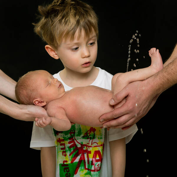 Ein unbekleidetes Baby wird am Po und an den Schultern von zwei paar Händen gehalten. Ein Bub steht dahinter und schaut fasziniert, wie das Baby in einem hohen Bogen pinkelt.