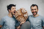 Zwei junge Männer mit grau-blauen Bäckerei-T-Shirts halten gemeinsam einen Laib Brot. einer der Männer ist gerade kurz davor, in den Laib zu beißen.