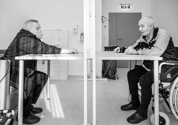 Zwei alte Männer sitzen sich gegenüber. Der rechte sitzt in einem Rollstuhl. links vorne ist ein zweiter Rollstuhl erkennbar. Der linke Mann trägt einen Mund-Nasen-Schutz. Auf dem Tisch zwischen den Beiden ist eine Schutzscheibe aufgebaut. Schwarz-Weiß.