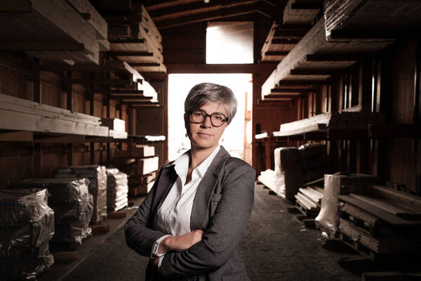 Eine Frau mit Brille steht mit verschränkten Armen in einer Lagerhalle.