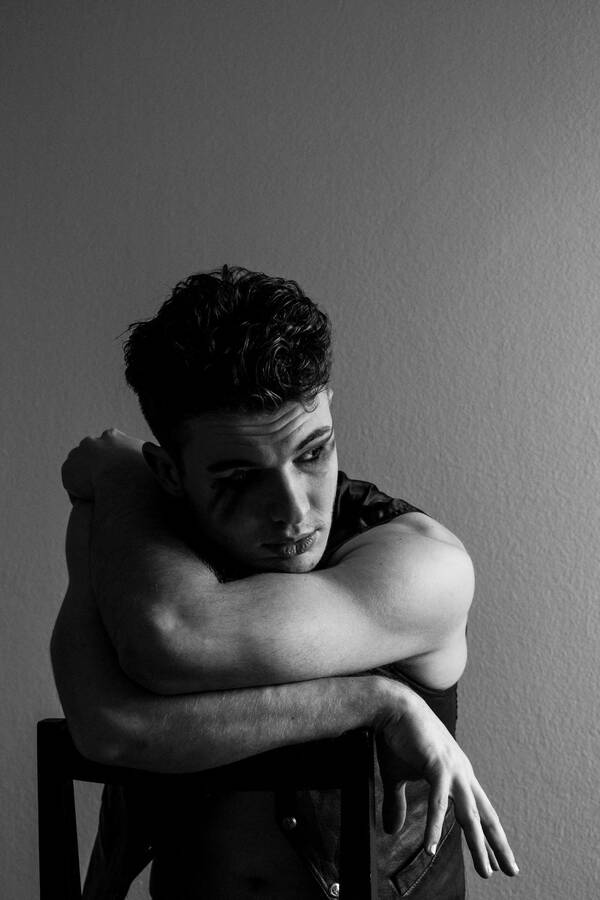 Portrait eines jungen Mannes, der rittlings auf einem Stuhl sitzt und die beiden muskulösen Arme auf die Lehne des Stuhles gelegt hat. Schwarz-Weiß.