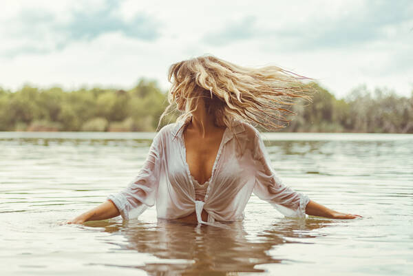 Eine Frau mit langen blonden Haaren in einer weißen Bluse steht bis zur Hüfte im Wasser eines sees und dreht den Kopf, so dass die Haare fliegen.