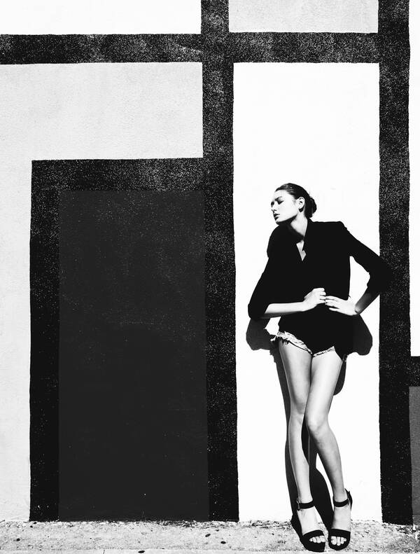Eine junge Frau in Hotpants und schwarzem Blazer steht in Modelpose vor einer weißen Wand, auf die mit schwarzer Farbe rechtwinklige geometrische Strukturen gemalt sind. Schwarz-Weiß.