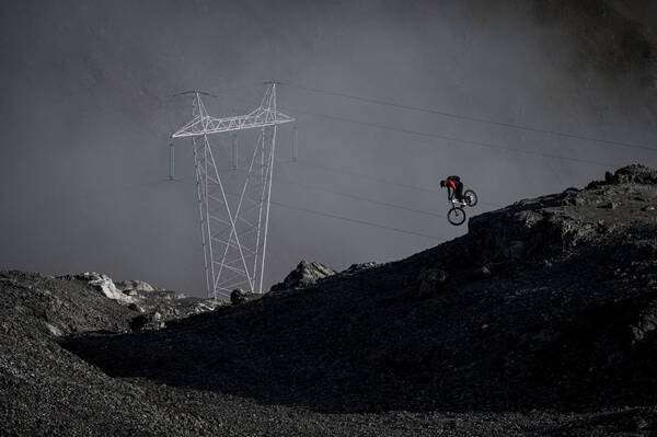 Ein Mountain-Biker springt über einen Felsblock im Gebirge, im Hintergrund ein doppelter Strommast.
