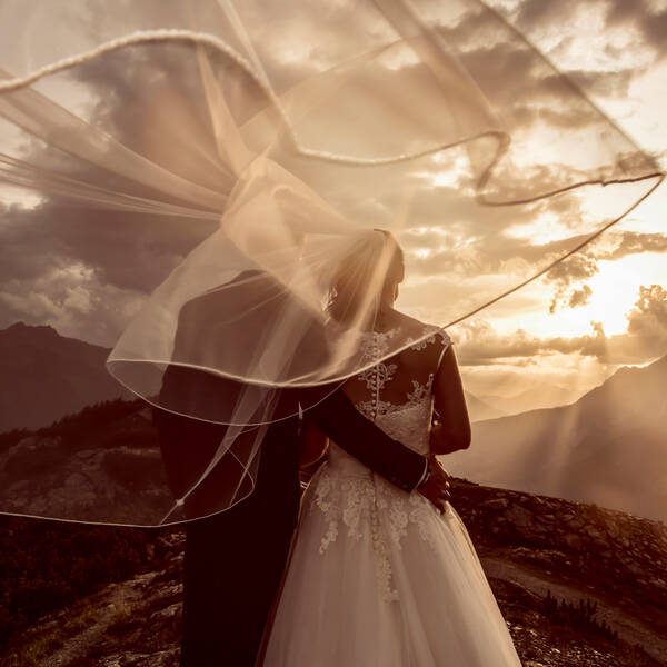 Ein Brautpaar im Abendlicht von hinten, er hat ihr den Arm um die Hüften gelegt. Der Schleier wird vom Wind über die obere Bildhälfte geweht. 