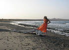 Frau in orangem Strandkleid und mit Strohhut an einem Strand, ein Fuß auf einem Stück Treibholz.