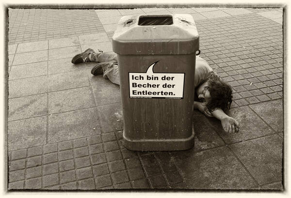 Ein Mann liegt hinter einem Abfallbehälter auf dem gepflasterten Boden. Auf dem Abfallbehälter steht in einer Sprechblase: 
