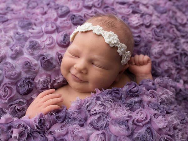 lächelndes Baby mit Blumenstirnband liegt in lila Blumen-Bettzeug.