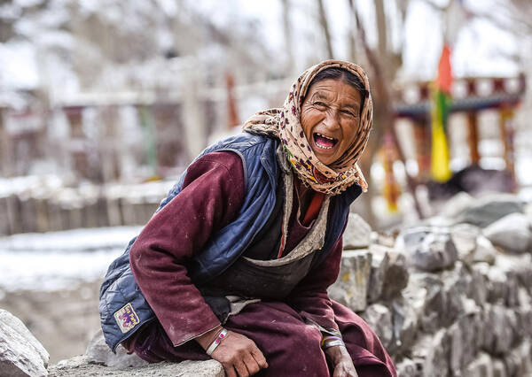 Eine alte Frau mit Kopftuch und ärmelloser Steppjacke sitzt auf einer winterlichen Steinmauer und Lacht.