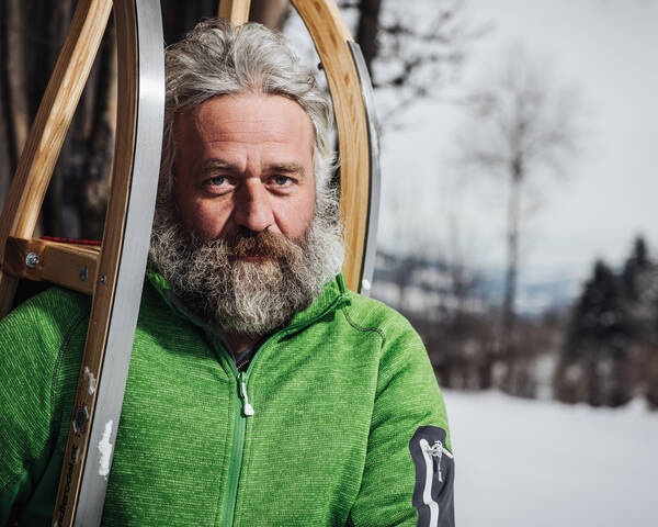 Ein weißhaariger, bärtiger Mann in grüner Fließjacke in einer Winterlandschaft trägt einen Schlitten auf der Schulter.