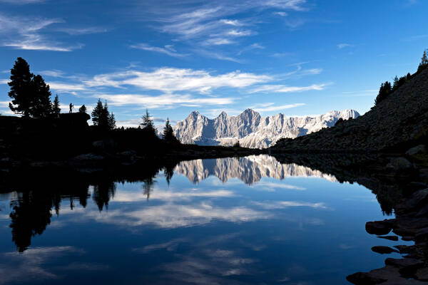 Panorama des Dachsteinmassives das sich im Spiegelsee auf der Reiteralm spiegelt. die Bäume im vordergrund sind nur als Silhouette wahrnehmbar. Teil dieser Silhouette ist auch ein Fotograf mit Stativ.