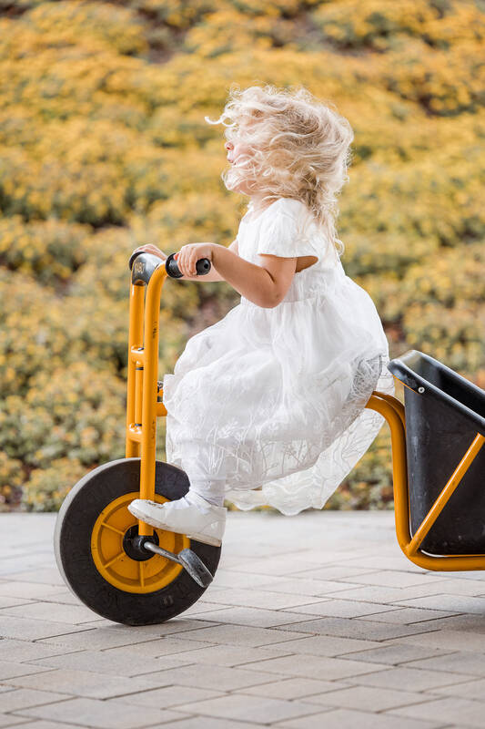 ein kleines Mädchen in einem weißen Kleid sitzt auf einem dottergelben Dreirad. Der Wind weht ihr von Hinten die blonden Locken ins Gesicht.