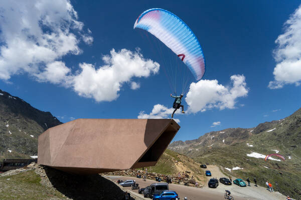 Ein Paragleiter startet vom Dach des Timmelsjoch-Museums. Im Hintergrund sieht man parktende Autos, Radfahrer, Touristen und andere Paragleiter.