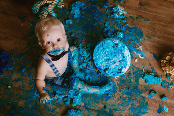 Ein Kleinkind mit nacktem Oberkörper und Mascherl sitzt auf einem Holzboden inmitten der rundum am Boden verteilten Überreste einer blau durchgefärbten Torte, auch die nackten Beine, Hände und Mund des Kindes sind mit Tortenresten verschmiert.