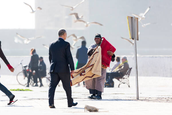 Ein Teppichhändler auf der Straße, Passanten und Möwen im Hintergrund.