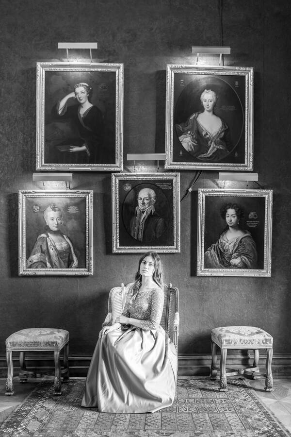 Eine Frau in elegantem Kleid sitzt in einem barocken Lehnstuhl unter historischen Gemälden – Eines zeigt einen Mann, die anderen Portraits zeigen Frauen.. Schwarz-Weiß.