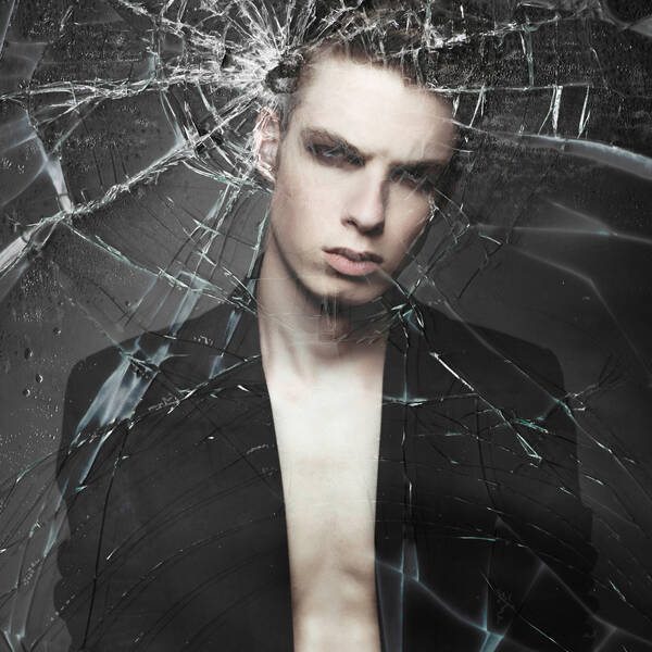 Portrait eines jungen Mannes in geöffnetem Hemd und strengem Blick durch eine zerschossene Glasscheibe fotografiert.