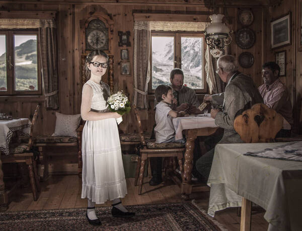 Ein Mädchen in weißem Kleid mit weißem Blumenstrauß in einer holzgetäfelten Almstube. Im Hintergund spielen vier Leute Karten, ein Bub, ein junger Mann, ein mittelalter Mann und ein alter Mann.