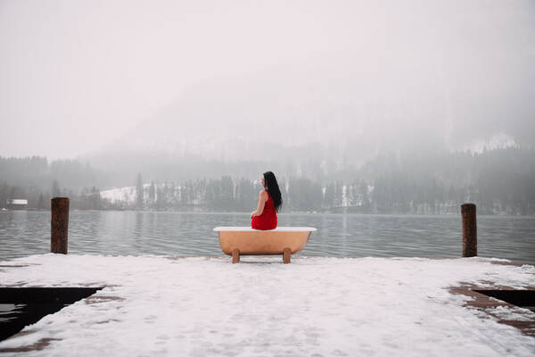 Eine Frau in rotem Kleid mit langen, schwarzen Haaren, sitzt auf einer godenen Badewanne auf einem verschneiten Steg mit Blick auf den nebeligen See.