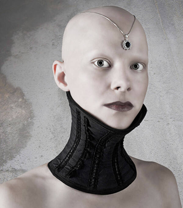 Portrait einer jungen, glatzköpfigen Frau mit schwarzer Halsmanschette. Auf dem Kopf trägt sie eine Kettemit einem edelsteinbesetzten Anhänger auf der Stirn.