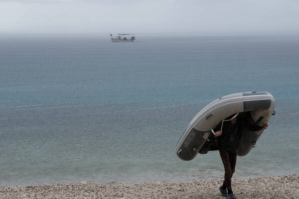 Ein Mann trägt an einem regnerischen Tag ein Schlauchboot vom Strand. im Hintergrund ein verankertes Segelschiff.