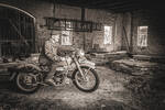 Ein alter Mann sitzt in einem Stadel auf einem Oldtimer-Motorrad. Schwarz-Weiß.