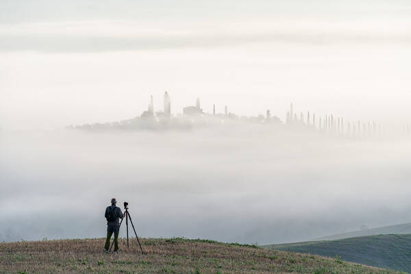 Ein Fotograf steht mit einem Kamerastativ vor einer nebeligen Landschaft. In der Ferne heben sich Zypressen und Gebäude aus dem Nebel ab.