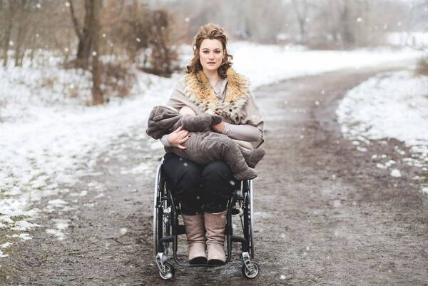 Eine Frau im Rollstuhl in Winterlandschaft stillt ihr Kind.