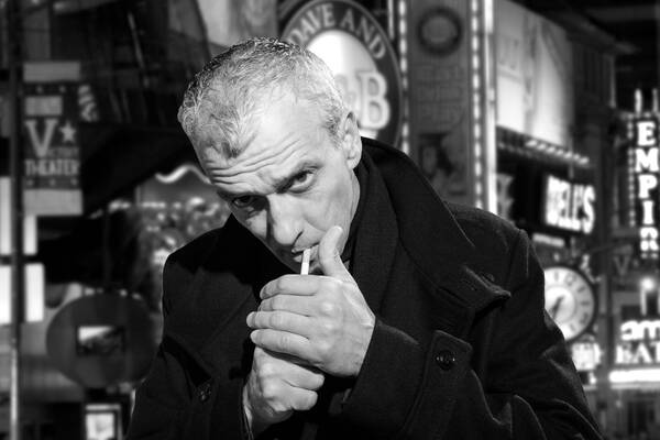 Ein Mann in schwarzem Mantel  zündet sich in einer Bar eine Zigarette an. Schwarz-Weiß.