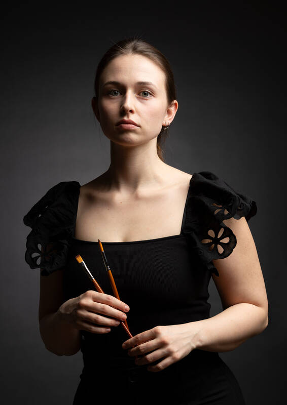 Portrait einer jungen Frau in schwarzem Kleid vor schwarzem Hintergrund. Sie hält zwei Pinsel in der Hand.