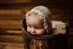 Ein Kleinkind mit weißer Spitzenhaube sitzt in einem alten hölzernen Kübel und nagt am Holz.