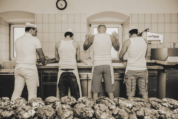 Vier Männer stehen in Bäcker-Arbeitskleidung an einer Arbeitsplatte beim Broteformen. im Vordergrund liegen auf einem Tisch gebackene Brote. Duotone. Schwarz-Weiß.