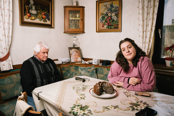 Eine junge Frau sitzt in einer traditionellen Stube mit einem alten Mann am Tisch. Im Hintergrund ist das Portrait einer alten Frau. Am Tisch steht ein Kuchen und es liegen Mobiltelefone und Fernsteuerungen am Tisch.