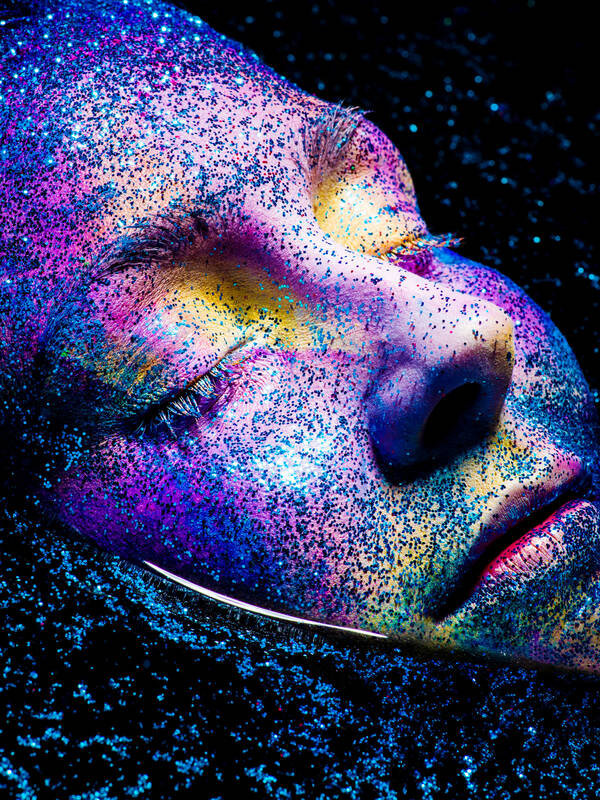 Close-up-Portrait einer Frau mit geschlossenen Augen, deren Gesicht in bunten Farben bemalt und mit Farbpigmenten bestreut ist. 