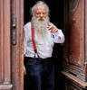 Portait eines älteren Mannes mit weißem Rauschebar, Zigarre und roten Hosenträgern. der Mann steht in einer geöffneten alten Eingangstüre.