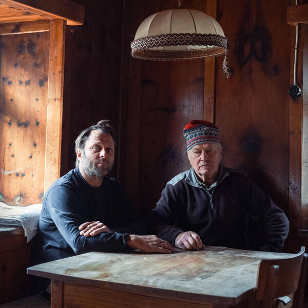 Zwei Männer sitzen an einem rustikalen Holztisch in einer alten Stube. Einer trägt eine Schimütze.